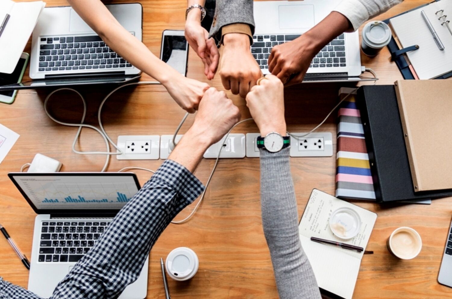 Grupo de pessoas cumprimentando com um soquinho em conjunto, sobre mesa com vários laptops e objetos de escritório